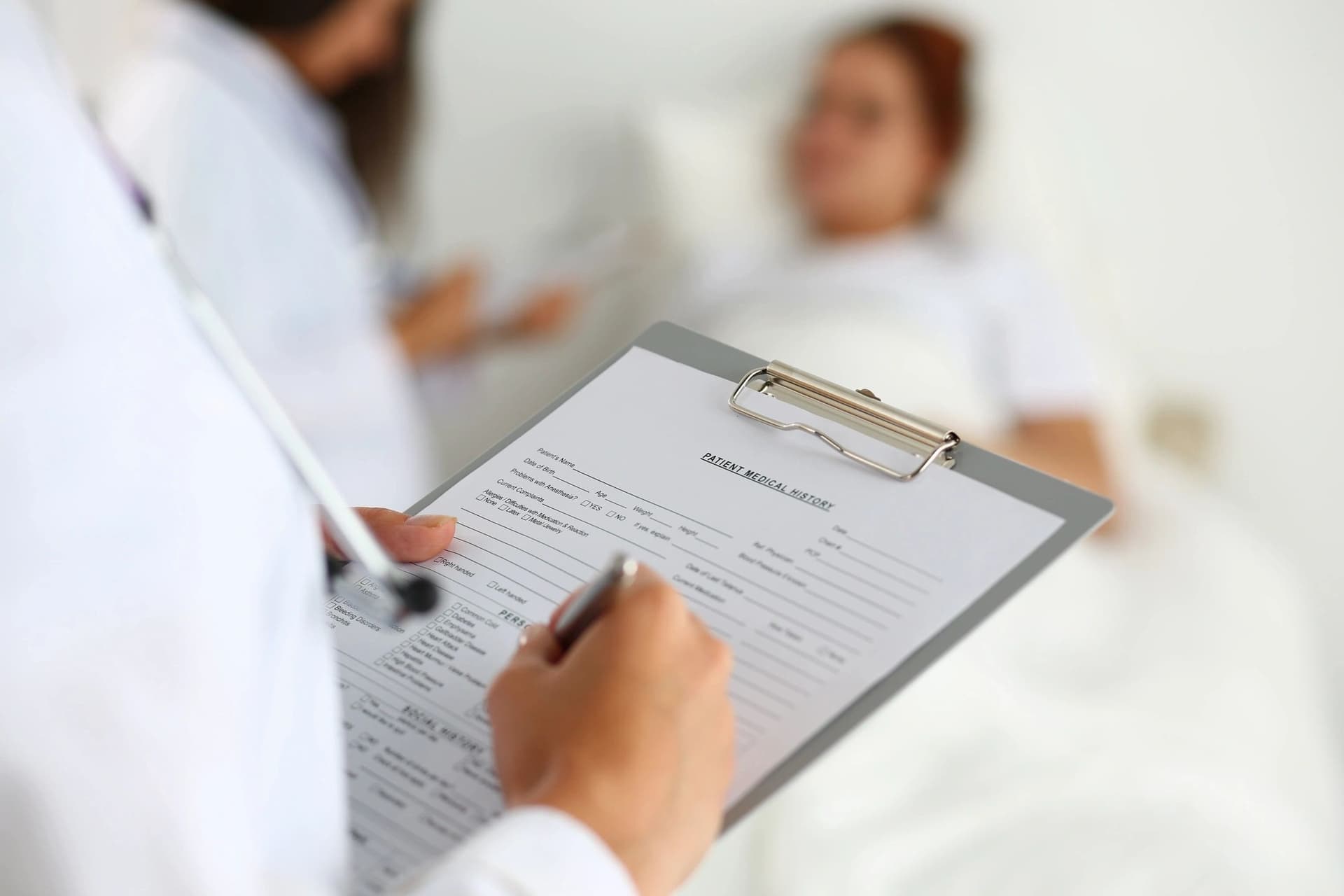 A clinician filling in a patient form on a clipboard beside a patient resting in bed