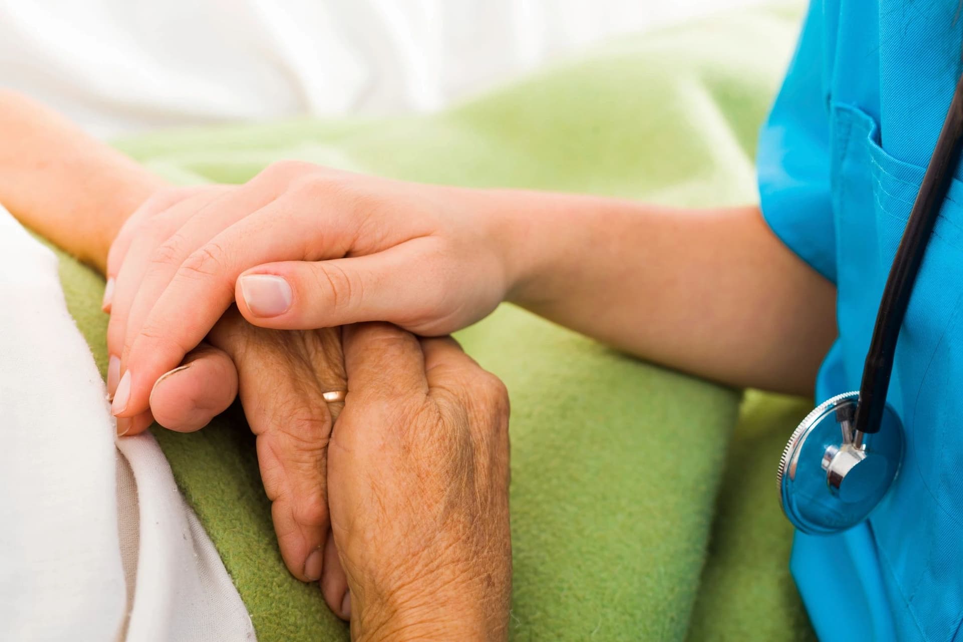 A nurse with a stethoscope holding an elderly patient’s hand resting on a blanket