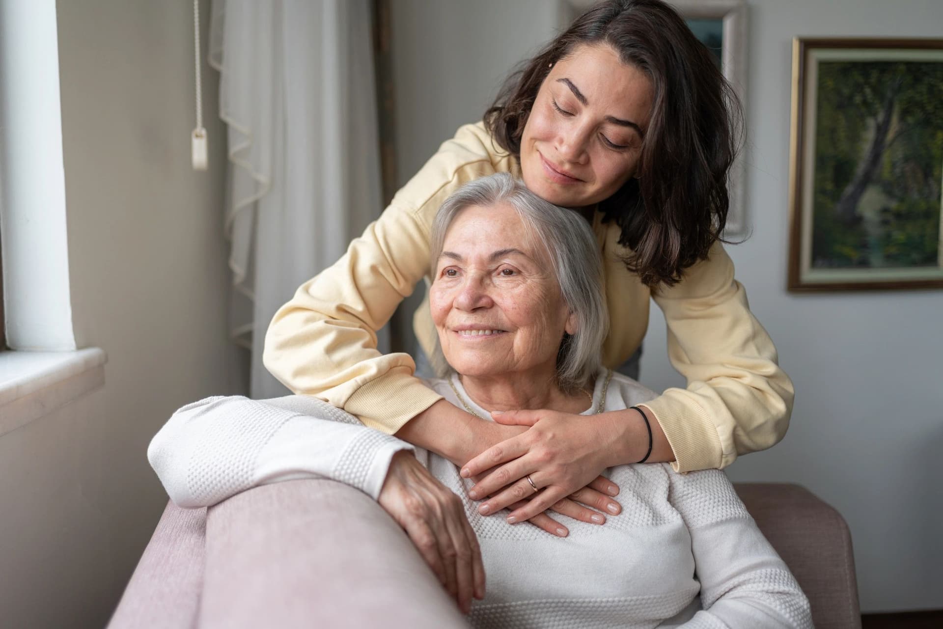 A smiling young woman hugging her elderly mother from behind on a sofa at home