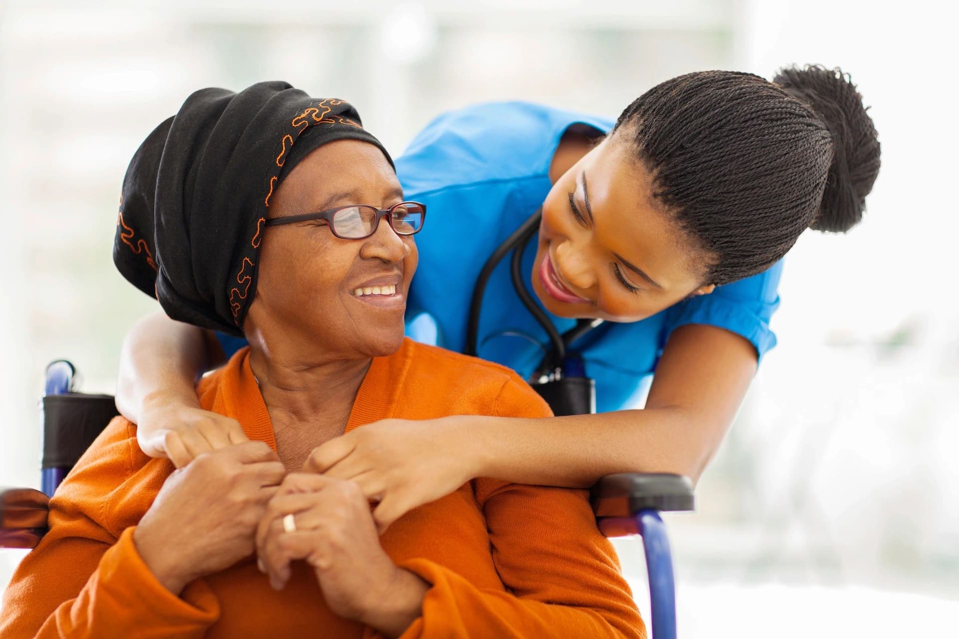 A caregiver in blue scrubs warmly embracing a smiling elderly woman in a wheelchair