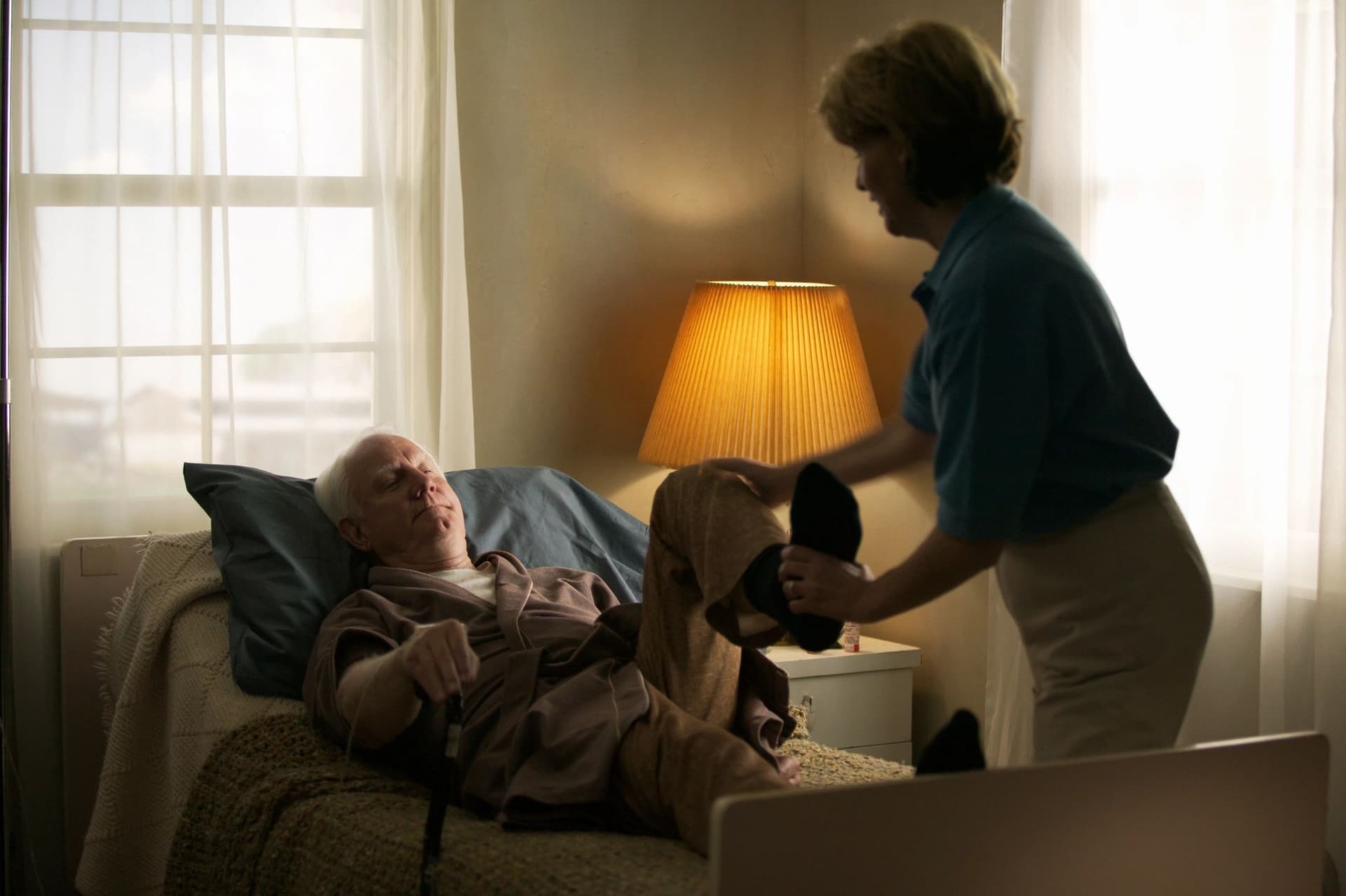 A caregiver helping an elderly man settle into bed in a softly lit bedroom