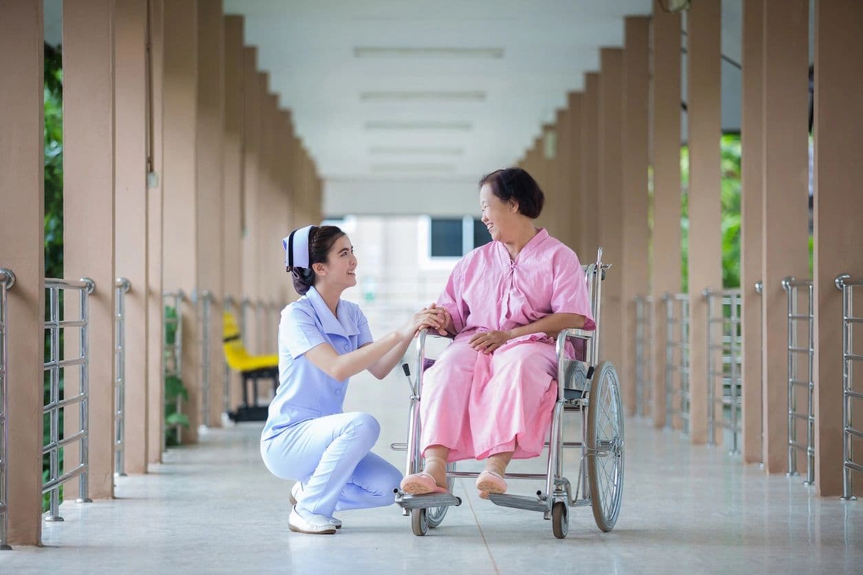 A nurse kneeling to talk with an elderly woman in a wheelchair in a bright corridor