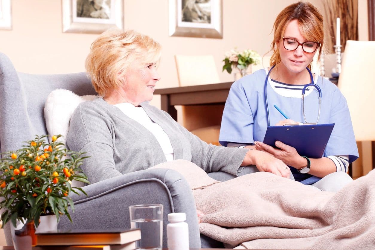 A nurse with a tablet talking with a smiling older woman seated in her living room